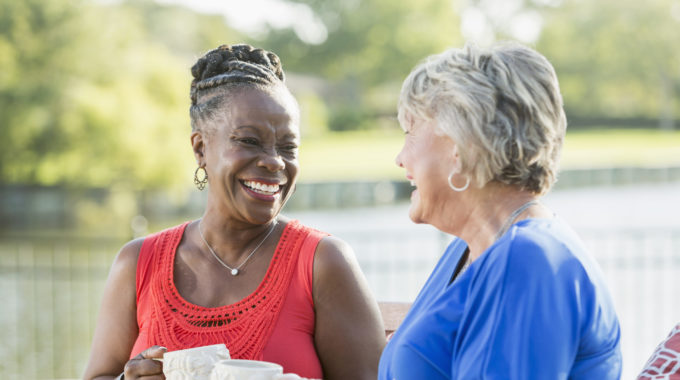 Two Senior Multi-ethnic Woman, One Caucasian And The Other African American, Sitting In Back Yard By Water Enjoying Each Other's Company. They Are Looking At Each Other, Laughing.