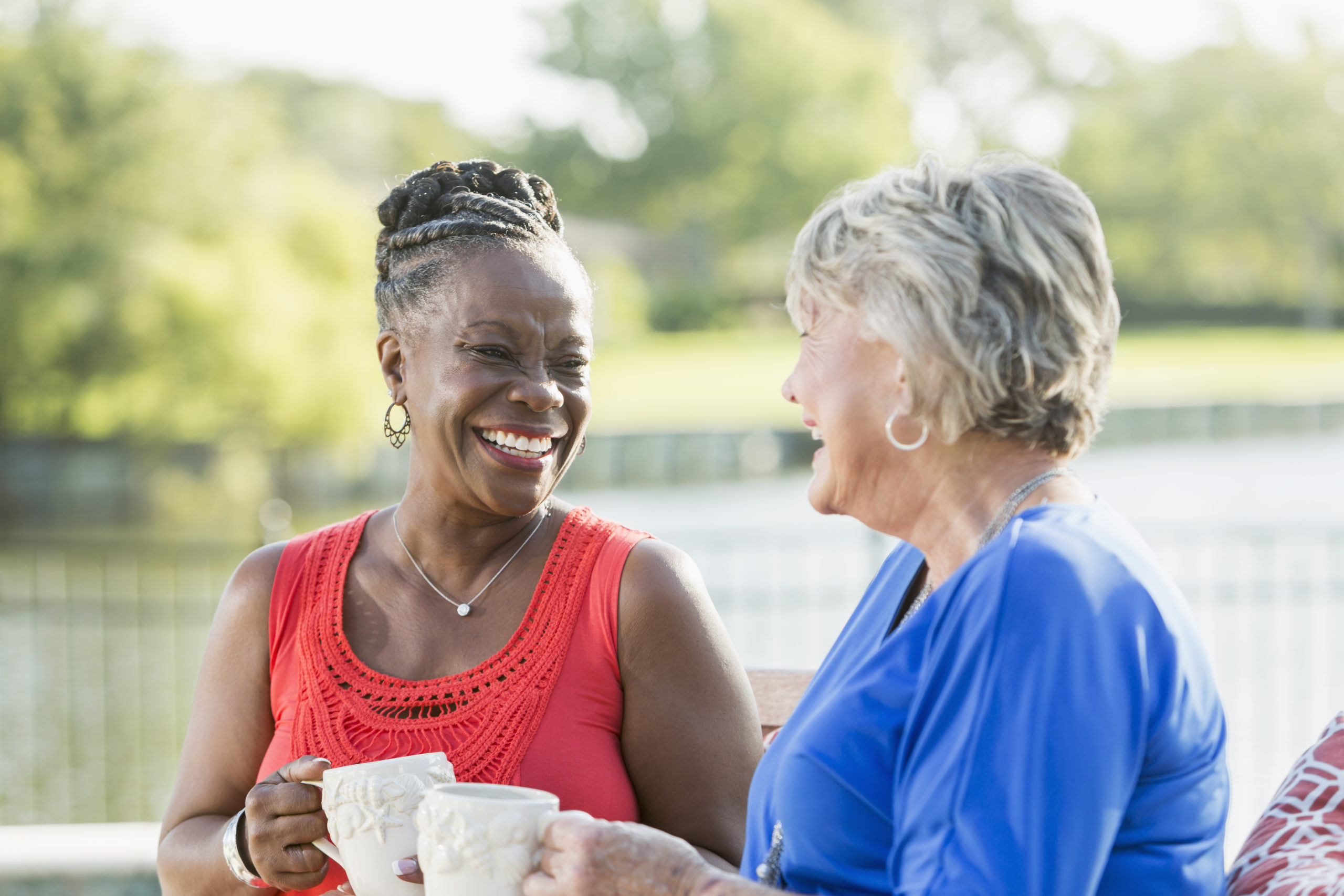 Two senior multi-ethnic woman, one Caucasian and the other African American, sitting in back yard by water enjoying each other's company. They are looking at each other, laughing.